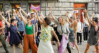 Viele Menschen stehen zusammen und strecken entschlossen die rechte Hand in die Höhe. Eine Frau hält eine lila Protest-Fahne in der Hand.