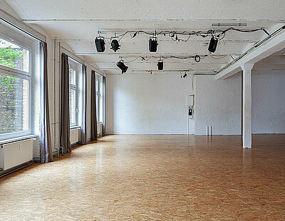Ein leeres Studio mit Holzboden. Auf der linken Seite sind drei große Fenster mit offenen grauen Vorhängen. Sechs kleinere Scheinwerfer hängen an der Decke.