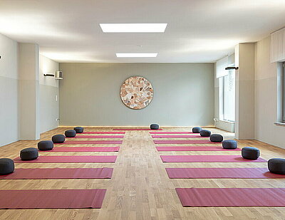 Zu sehen ist das Studio im Cornus Berlin. Auf dem Holzboden sind mehrere rote Yogamatten ausgerollt. Darauf liegen graue Kissen.