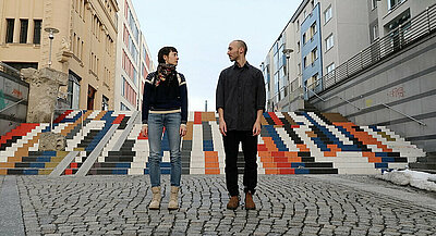 Nitsan Margaliot und Sasha Portyannikova stehen auf einem gepflasterten Fußweg und schauen sich an. Hinter ihnen eine große bunte Treppe.