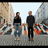 Nitsan Margaliot und Sasha Portyannikova stehen auf einem gepflasterten Fußweg und schauen sich an. Hinter ihnen eine große bunte Treppe.