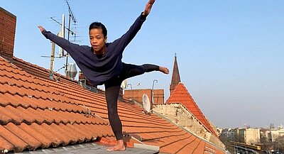 Zaratiana Randrianantenaina auf dem Dach ihres Hauses. Sie tanzt das Solo aus „gefaltet“ von Sasha Waltz. Sie hat beide Arme von sich gestreckt und steht in einer Standwaage.