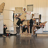 Eine Aufnahme der Performance "Zusammen bauen" von Gabi dan Droste. Ein Bühnenelement aus verschienenhölzernen Kisten, Platten und Rahmen - drumherum sitzen und stehen neun Performende.