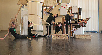 Eine Aufnahme der Performance "Zusammen bauen" von Gabi dan Droste. Ein Bühnenelement aus verschienenhölzernen Kisten, Platten und Rahmen - drumherum sitzen und stehen neun Performende.