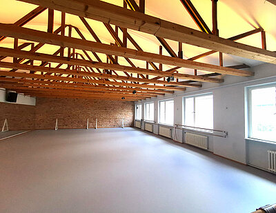 Ein Studio mit weißem Boden und verspiegelter Wand links. Gegenüber sind vier große Fenster. Oben sieht man mehrere Dachbalken aus Holz.