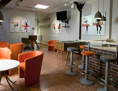 Zu sehen ist die X-Step-Cafeteria mit gefliestem Boden einer kleinen Bar mit Stehhockern rechts, zwei Sitzecken links und einem Tischkicker im Hintergrund.